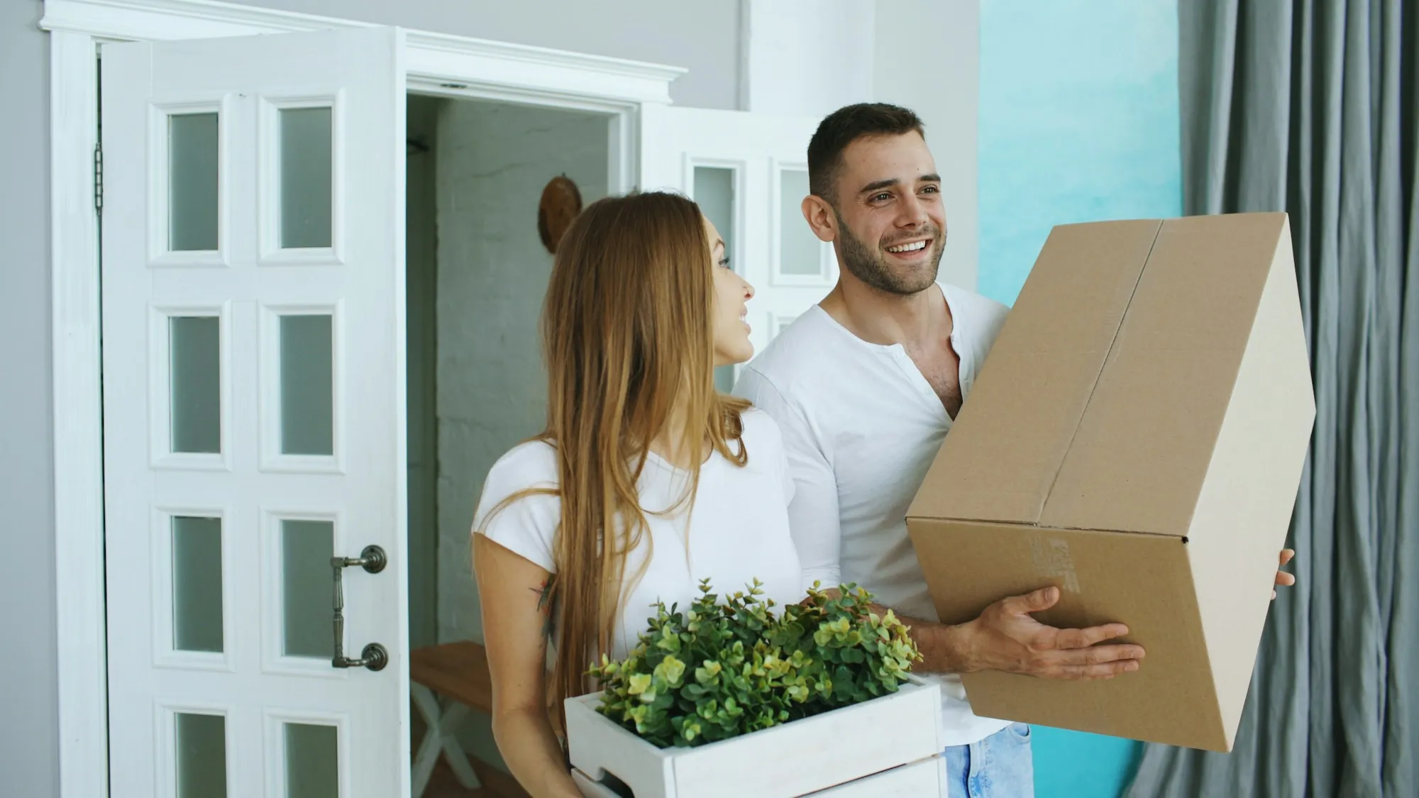 Smiling couple carrying a moving box and a potted plant into their home